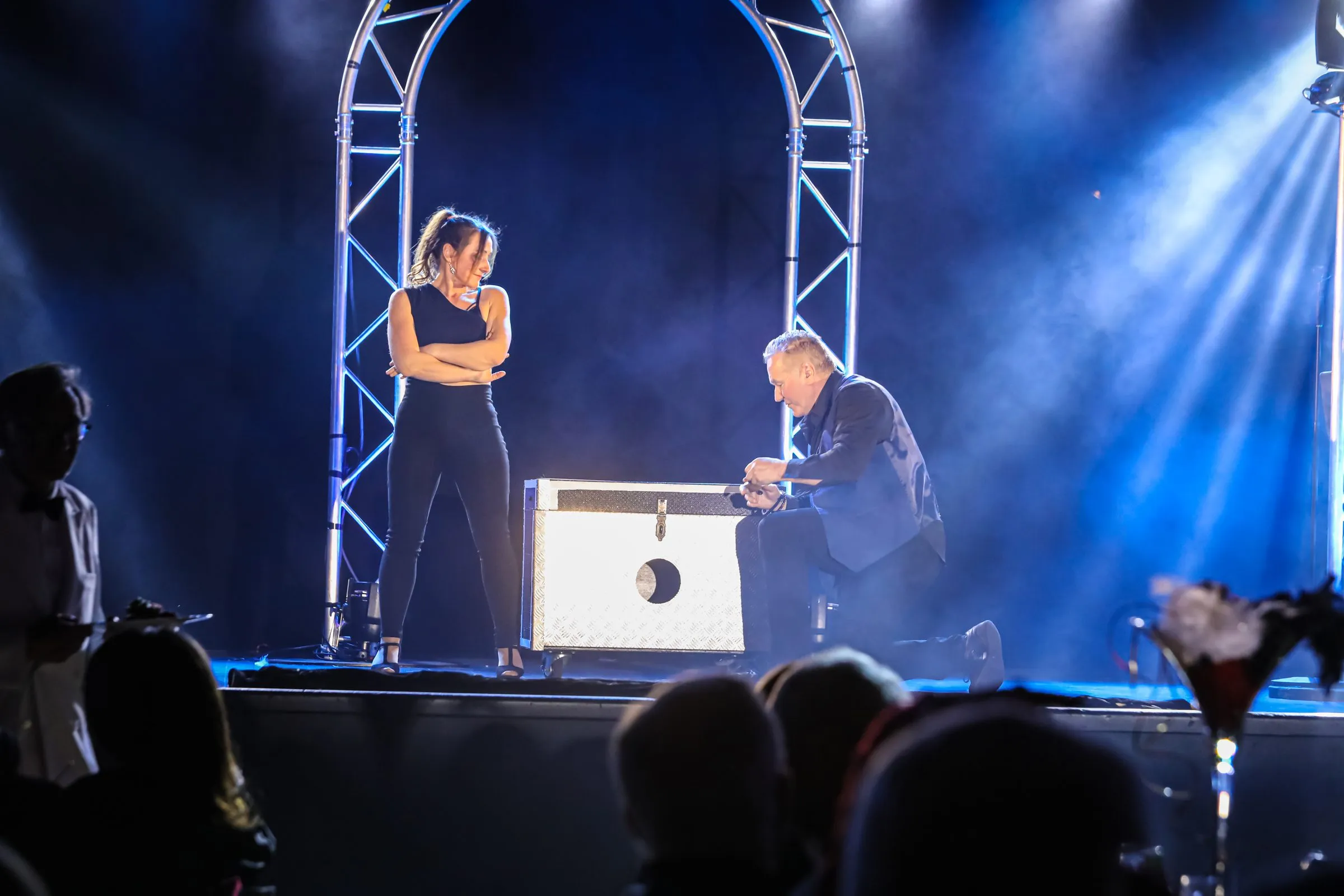 Mickaël Ross agenouillé devant une caisse lors d'une grande illusion, sous une arche lumineuse